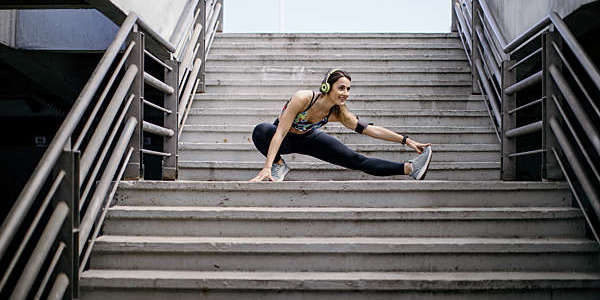Young woman wearing headphones, crouched halfway up a flight of steps, performs stretching exercises before her run.