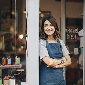 Female owner of a delicatessen smiles confidently as she stands in the doorway of her establishment.