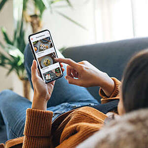 Over-the-shoulder view of a young woman slumped on the sofa as she selects food from a mobile app menu.