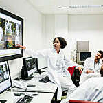 A clinical registrar reviewing a patients test results, looking at some information on a large monitor with some other doctors.