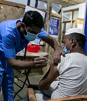 Male nurse in blue uniform administers the COVID-19 vaccine to a Rohingya refugee seen from behind.